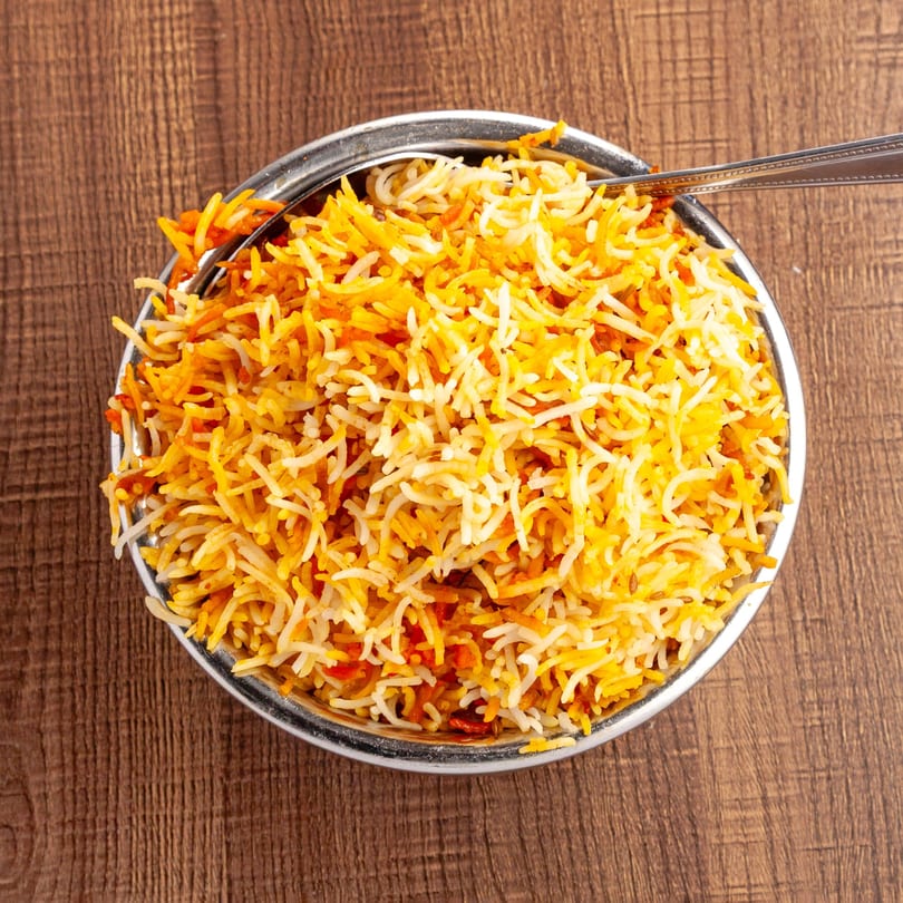 A metal bowl of vibrant orange and white Pakistani biryani rice with a spoon, on a wooden table.